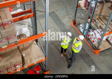 Directly above view of coworkers in green vests and hardhats discussing new supply at warehouse while looking at boxes on shelves Stock Photo