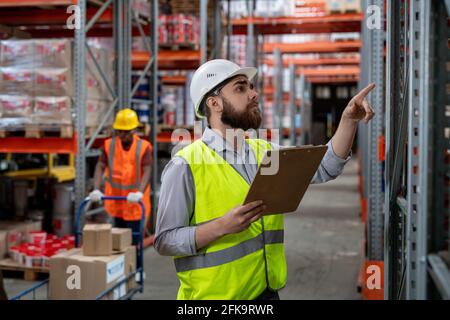 Serious pensive bearded warehouse manager in white hardhat and green vest holding clipboard and counting boxes while inspecting goods Stock Photo
