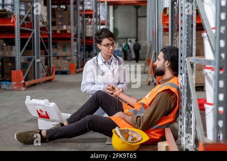 Warehouse nurse with stethoscope around neck sitting on floor and checking pulse of mover dropped on floor Stock Photo