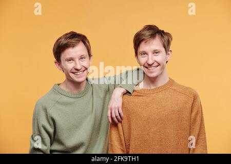 Portrait of happy young man in casual sweater leaning on shoulder of twin brother against orange background Stock Photo