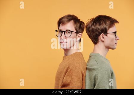 Portrait of content young man in mustard sweater and glasses standing back to back with twin brother against orange background Stock Photo