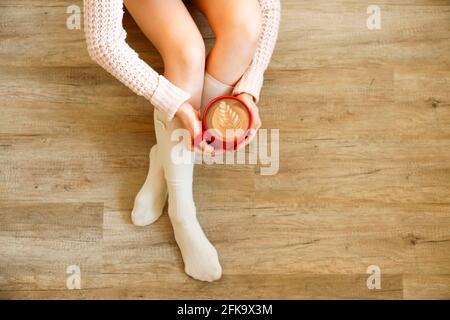 Young woman drinking cappuccino coffee and sitting on the wooden floor. Top view of female legs in warm white knee high socks. Comfort winter holidays