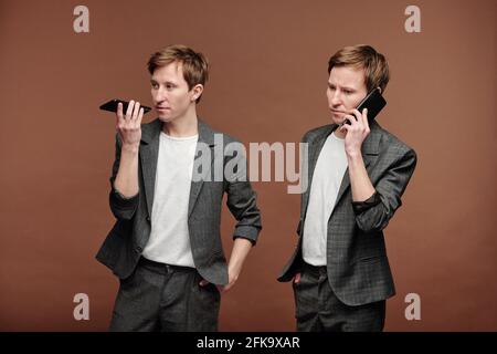 Confident young man in suit recording voice message while his twin brother listening to it on phone Stock Photo