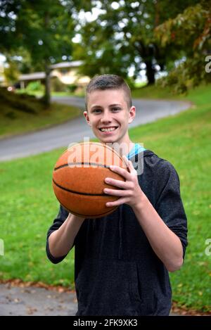 Young teen practices basketball in his yard.  He is holding a ball and smiling at the camera. Stock Photo