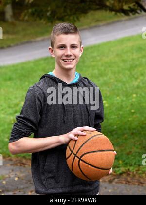 Young teen stands, in his backyard, holding a well worn basketball.  He is looking at the camera and smiling.  He has on a hoodie sweatshirt with the Stock Photo