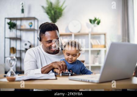 Casual african businessman in headset having working meeting on laptop while nursing his baby son at home. Concept of technology and freelance. Stock Photo