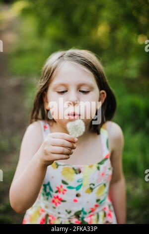 Young girl blowing on a dandelion surrounded by trees and other greenery Stock Photo