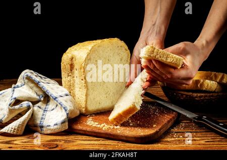 Female hands breaking a piece of fresh white homemade bread that stands on a wooden table. Homemade cakes, pastries. Stock Photo