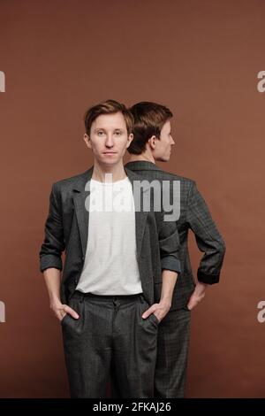Portrait of content confident young man in gray suit standing back to back with brother, brown background Stock Photo
