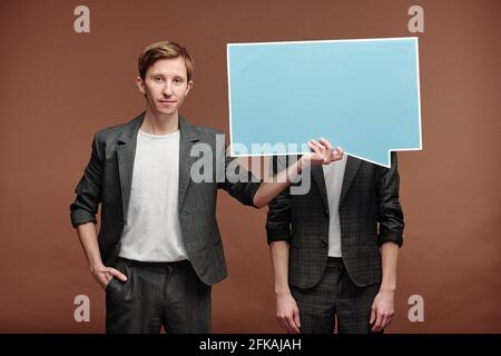 Portrait of content young man in smart casual suit hiding face of man behind speech bubble tag against brown background Stock Photo