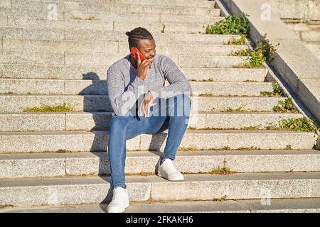 Happy black man talking on smart phone sitting on stairs. Young man in jeans and grey sweater. Concept of communication. High quality photo Stock Photo