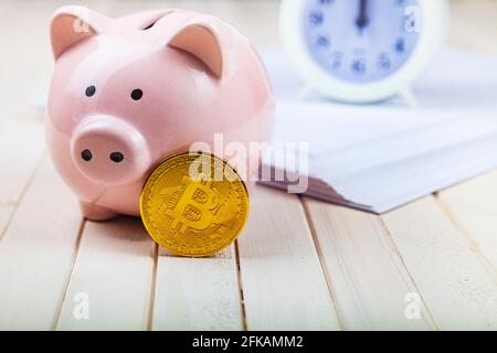 Pig piggy bank and bitcoin, clock and stack of papers on the background. Stock Photo
