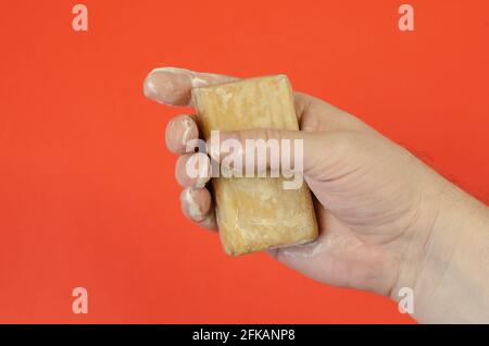 Hand and wet laundry soap on a red background. The man is holding a rectangular bar of brown soap. The palm is covered with soapy foam. Stock Photo