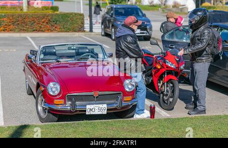 Two old man talking in front of a car in a street parking lot. One with the bike and one with the vintage red car. Stock Photo