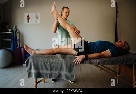 Female physiotherapist stretching hamstring of elderly man lying on massage bed in exercise room. Stock Photo