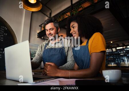 Manager of coffee shop training intern showing menu on laptop while drinking hot coffee in cafe  Stock Photo