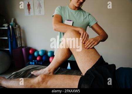 Female physiotherapist pinching nerves of elderly male patients knee while lying n on massage bed in pilates room. Stock Photo