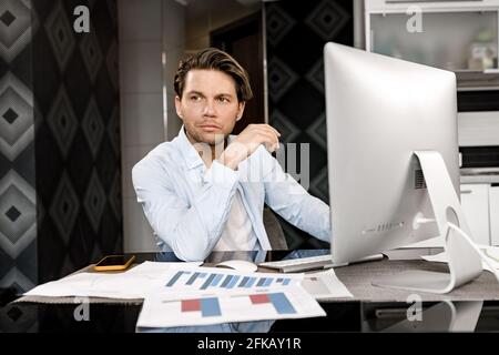 Pensive male freelancer working on new project, sitting at the desk in home office. Handsome young businessman or student lost in thoughts, developing Stock Photo