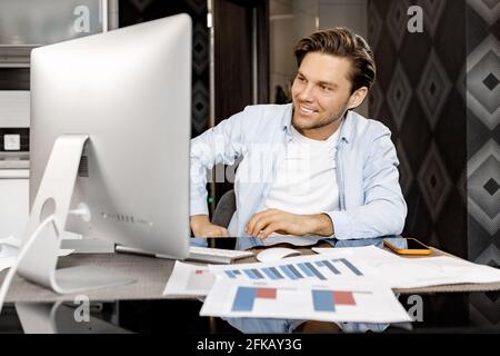 Handsome young businessman is having a video meeting with colleagues, sitting at the desk, using computer. Happy stylish male freelancer talking with Stock Photo