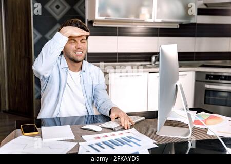 Caucasian male freelancer is sitting at the desk, having a terrible headache. Tired young businessman is using computer, learning financial graphs for Stock Photo