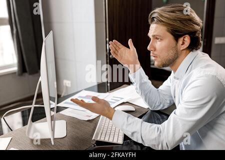 Serious male employee is sitting at the desk in home office, using computer, having a video conference. Focused young businessman talking with Stock Photo