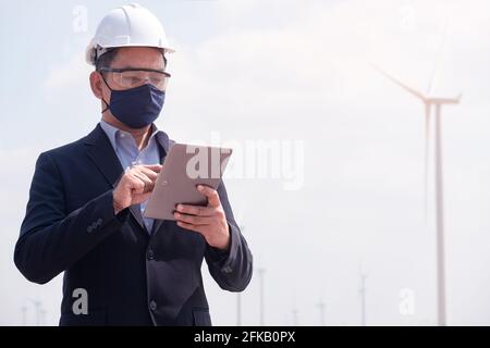 Successful engineer windmills manager wearing face mask  and using tablet with wind turbine in background Stock Photo