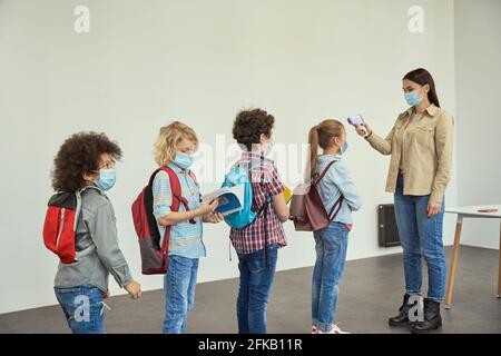 New rules. Modern young female teacher measuring temperature, screening kids with digital thermometer while students coming to school Stock Photo