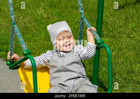 Little Caucasian girl 2 years old swinging on a swing and laughing cheerfully. Cheerful kid in a gray hat plays on the playground, close-up portrait Stock Photo