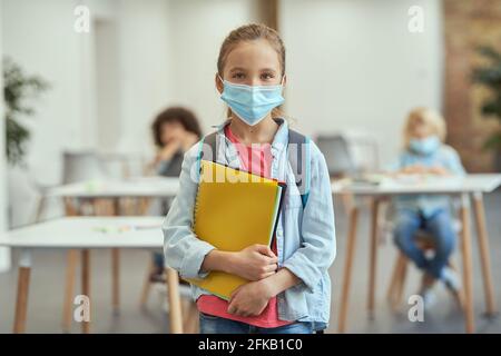 Portrait of adorable little school girl wearing mask to prevent the spread of Covid19 looking at camera, holding textbooks while posing in a classroom Stock Photo