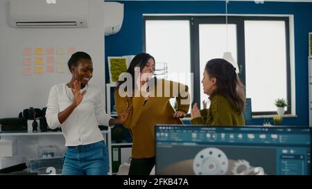 Enthusiastic diverse engineers women dancing in company office celebrating success of project, singing and having fun during working hours. Excited multiethnic engineers using pc showing cad software Stock Photo