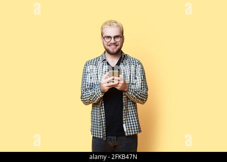 Blonde bearded man with glasses is smiling at camera while chatting on a mobile posing on a yellow studio wall Stock Photo