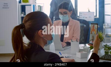 Back view of freelancer man with face mask talking on videocall meeting with remotely collegues using phone. Business woman working in new normal company office during covid19 pandemic Stock Photo