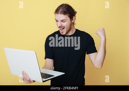 Caucasian man with long hair and beard is gesturing the yes sign posing on a yellow studio wall holding a laptop Stock Photo