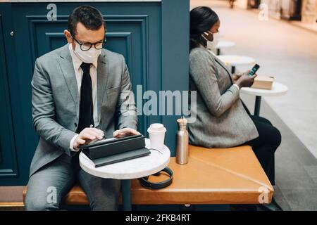 Male entrepreneur using digital tablet while female colleague with smart phone in cafe during pandemic Stock Photo