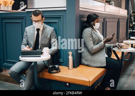 Businessman using digital tablet while female colleague with smart phone in cafe during COVID-19 Stock Photo