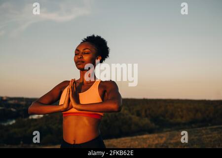 Young sportswoman meditating against sky during sunset Stock Photo