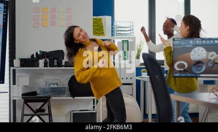 Happy diverse businesswomen dancing in company office celebrating success of project, singing and having fun during working hours. Excited multiethnic engineers using computer showing cad software Stock Photo