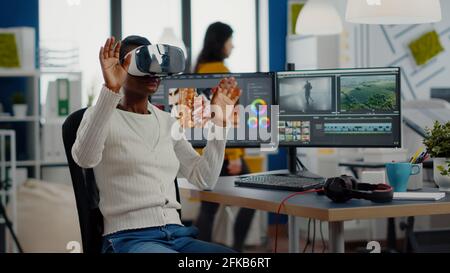 African video editor experiencing VR goggles, gesturing, editing video film montage working with footage and sound on computer with two displays. Videographer processing movie in creative agency Stock Photo