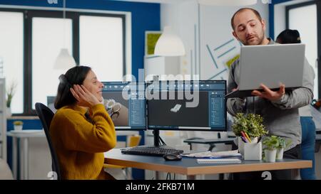 Industrial designer discussing with woman engineer while working in CAD program, designing new components, comparing 3D Prototype with project from laptop. Workteam using computer with two monitors Stock Photo