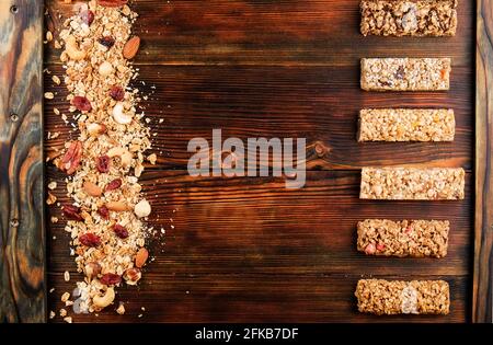 Variety of granola energy bars in row with scattered mixed nuts, cereals, cranberry on brown wood textured table background. Healthy nutritious vegan Stock Photo