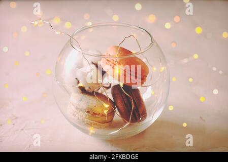 Stylish composition of French macaroons in glass bowl, cotton boll & Christmas lights, white stucco plaster textured table. Happy valentine Birthday w Stock Photo