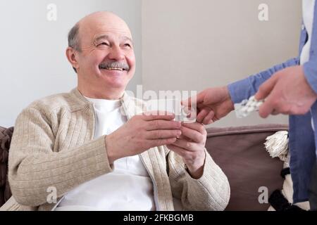 Elderly man sitting at home taking a glass of water from his son. Stock Photo