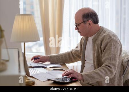 Senior man sitting with paperwork and using calculator while counting money Stock Photo