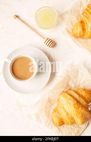 Cup of coffee with milk, tasty freshly baked french croissants and sweet honey jar. Healthy light breakfast on white stucco plaster textured table. Cl Stock Photo