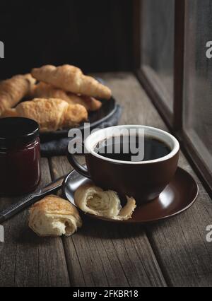 Hot cup of coffee with croissants on a rustic wooden table next to window Stock Photo