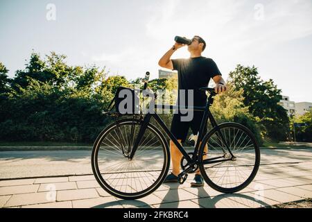 Sportsman drinking water while standing on footpath against sky on sunny day