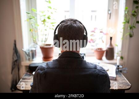 Rear view of man with headphones working on laptop at home during pandemic Stock Photo