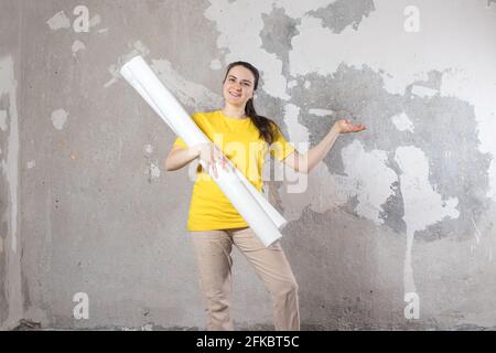 A woman in a bright yellow T-shirt holds 2 rolls of white wallpaper against the old wall. Repair work, wallpapering. With a place for text to copy spa Stock Photo