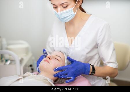 Face peeling and treatment at the beautician parlor. Woman cosmetologist applies a foam to the clients face with a hands. Preparing the teen girls face for the carboxytherapy procedure Stock Photo