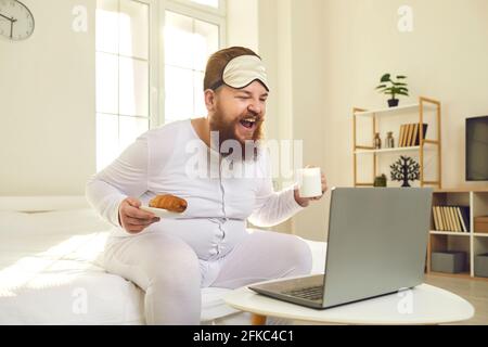 Man sitting on bed, enjoying morning coffee, watching funny movie on laptop and laughing Stock Photo